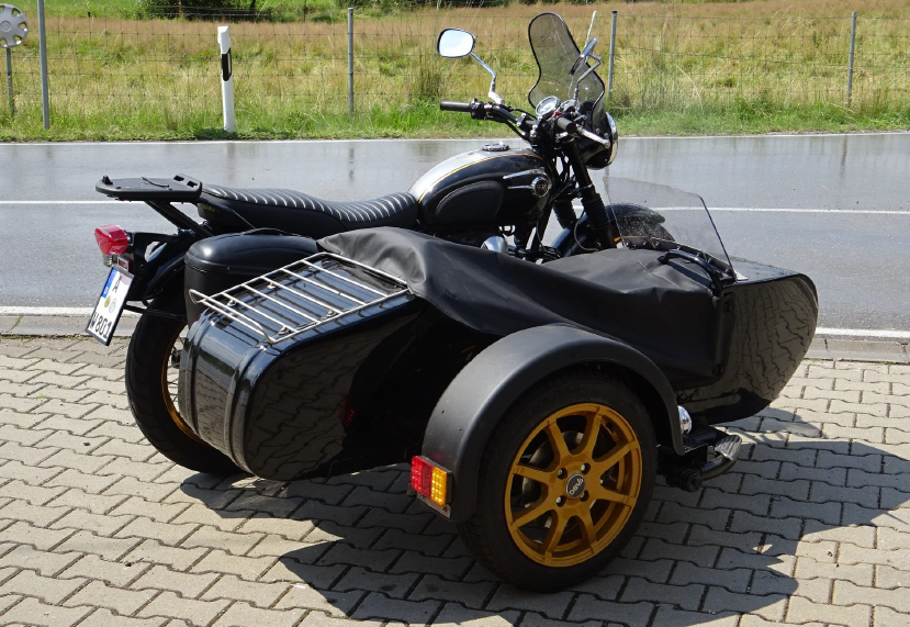Ein Motorrad mit Beiwagen, geparkt auf einer Kopfsteinpflasterstraße, umgeben von Grün und malerischen Gebäuden.