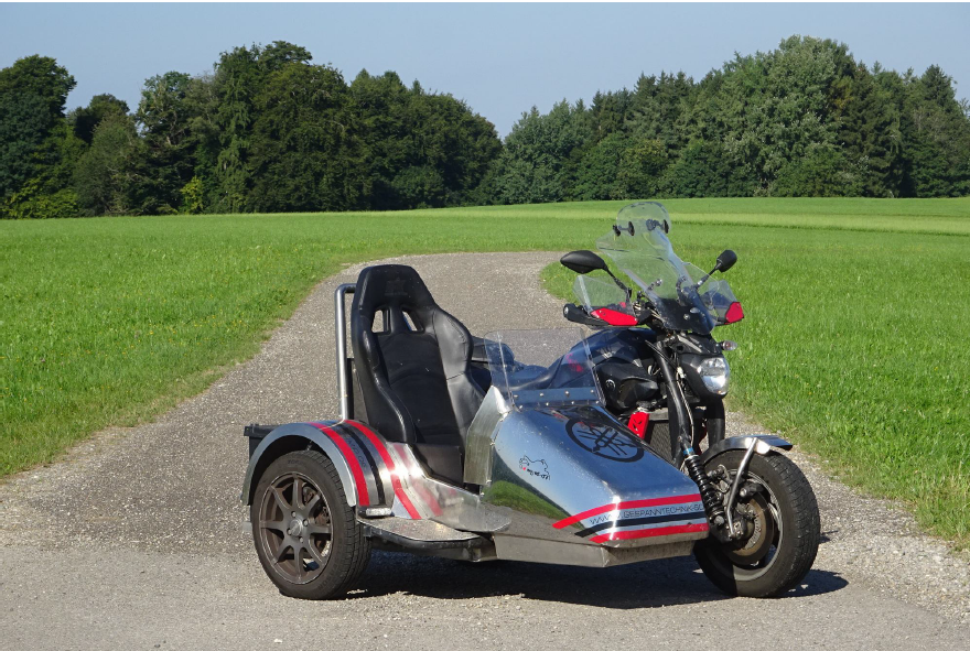 Ein Motorrad mit Beiwagen, geparkt auf einer asphaltierten Straße, umgeben von Grün und einem klaren blauen Himmel.